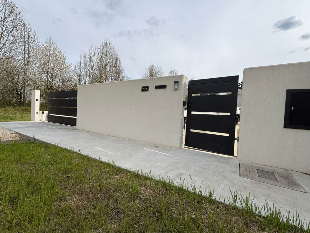 Cancello carraio scorrevole dal design moderno, realizzato in metallo nero a fasce orizzontali, inserito in un muretto di recinzione in cemento liscio Vista 3 - 2A Carpenteria