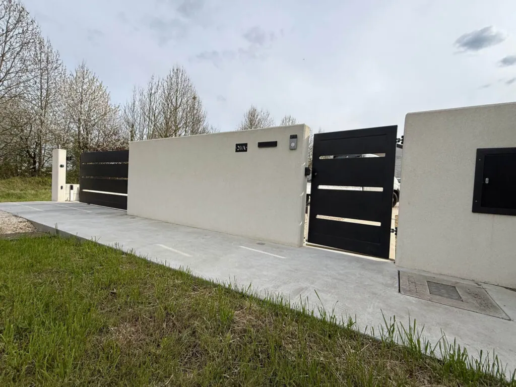 Cancello carraio scorrevole dal design moderno, realizzato in metallo nero a fasce orizzontali, inserito in un muretto di recinzione in cemento liscio Vista 3 - 2A Carpenteria