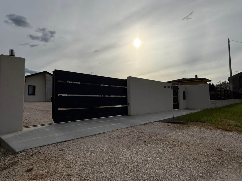 Cancello carraio scorrevole dal design moderno, realizzato in metallo nero a fasce orizzontali, inserito in un muretto di recinzione in cemento liscio Vista 2 - 2A Carpenteria
