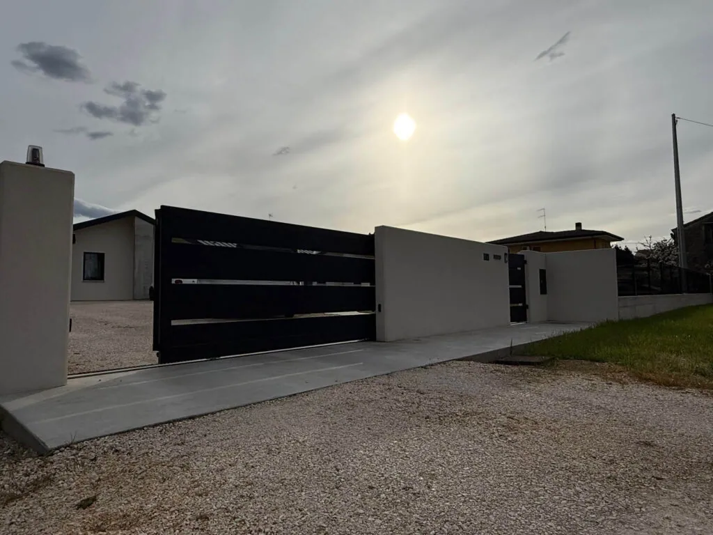 Cancello carraio scorrevole dal design moderno, realizzato in metallo nero a fasce orizzontali, inserito in un muretto di recinzione in cemento liscio Vista 2 - 2A Carpenteria