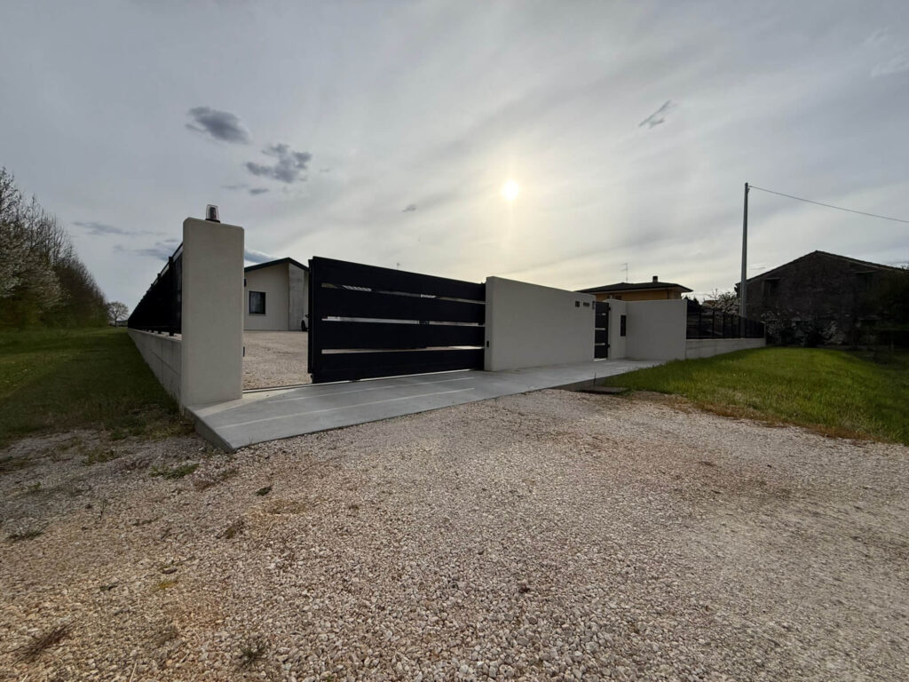 Cancello carraio scorrevole dal design moderno, realizzato in metallo nero a fasce orizzontali, inserito in un muretto di recinzione in cemento liscio - 2A Carpenteria