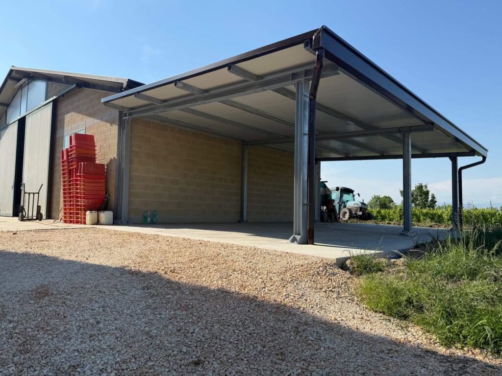 Grande tettoia metallica addossata a un edificio agricolo, realizzata con una solida struttura portante in acciaio e copertura in pannelli coibentati, adibita a rimessa per macchinari. Progettazione, realizzazione e posa a cura di Carpenteria 2A a Treviso, operante in tutto il Veneto - Vista 4.