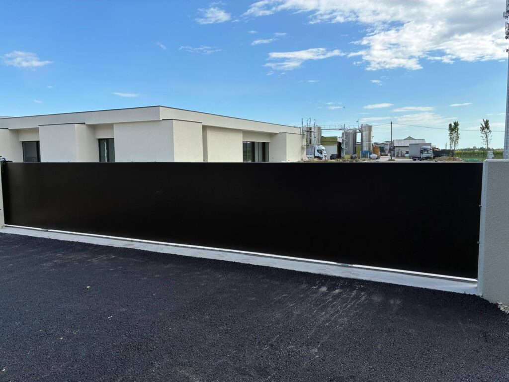 Lungo cancello carraio scorrevole dal design minimale, realizzato con pannelli ciechi in lamiera metallica nera per oscurare completamente la vista dall'esterno - Vista2