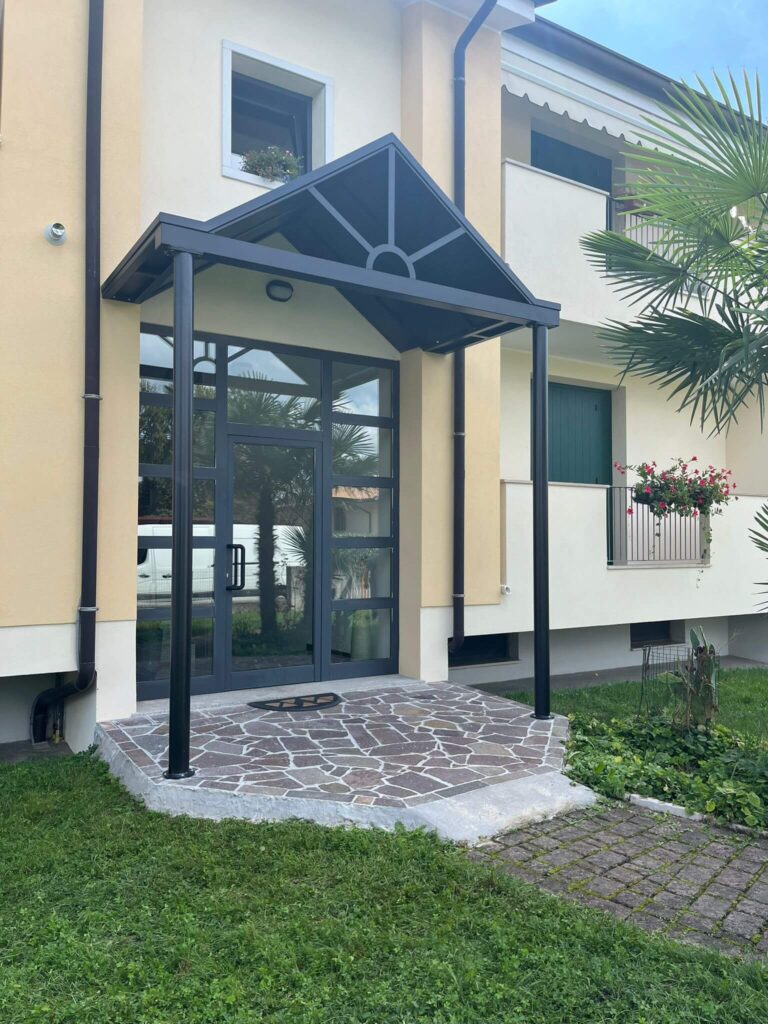 Elegante tettoia metallica a due falde posta a protezione del portoncino d'ingresso di un'abitazione, sorretta da due pilastri e impreziosita da un motivo decorativo a raggiera sul frontone. Struttura realizzata su misura da Carpenteria 2A a Treviso, Veneto.