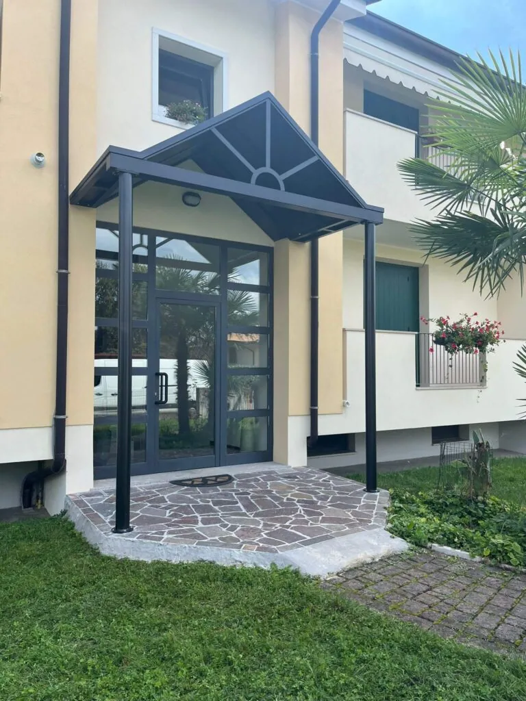 Elegante tettoia metallica a due falde posta a protezione del portoncino d'ingresso di un'abitazione, sorretta da due pilastri e impreziosita da un motivo decorativo a raggiera sul frontone. Struttura realizzata su misura da Carpenteria 2A a Treviso, Veneto.