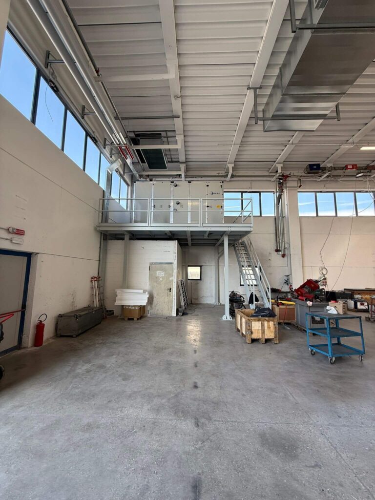 Vista frontale di un solido soppalco industriale in metallo verniciato chiaro, con scala di servizio e ringhiere a norma, a supporto di un'unità di condizionamento. Progettazione, realizzazione e posa a cura di Carpenteria 2A, operante in tutto il Veneto.