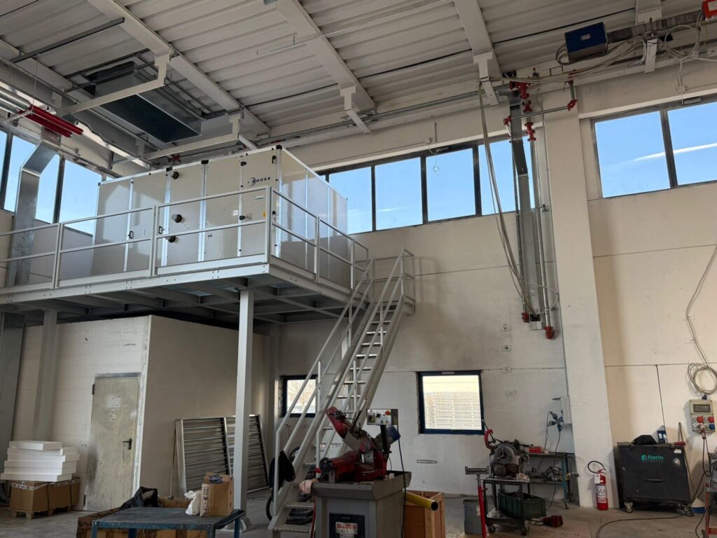 Struttura metallica portante a uso soppalco industriale, dotata di parapetto di sicurezza e scala d'accesso, progettata per sostenere un grande impianto di aerazione all'interno di un capannone. Opera di carpenteria pesante realizzata da Carpenteria 2A a Treviso, Veneto - Vista 2 - soppalchi in ferro.