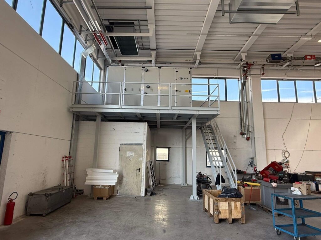 Struttura metallica portante a uso soppalco industriale, dotata di parapetto di sicurezza e scala d'accesso, progettata per sostenere un grande impianto di aerazione all'interno di un capannone. Opera di carpenteria pesante realizzata da azienda 2A a Treviso, Veneto.