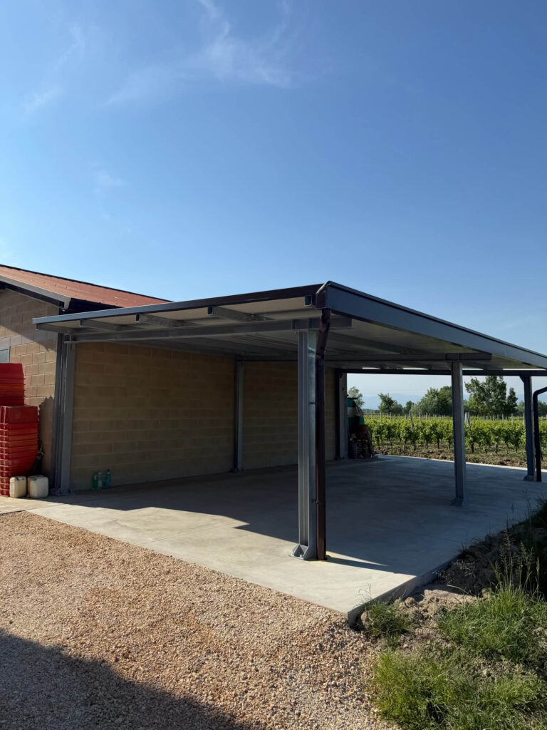 Grande tettoia metallica addossata a un edificio agricolo, realizzata con una solida struttura portante in acciaio e copertura in pannelli coibentati, adibita a rimessa per macchinari. Progettazione, realizzazione e posa a cura di Carpenteria 2A a Treviso, operante in tutto il Veneto - Vista 3.
