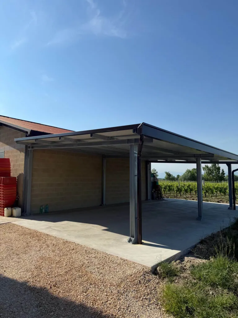Grande tettoia metallica addossata a un edificio agricolo, realizzata con una solida struttura portante in acciaio e copertura in pannelli coibentati, adibita a rimessa per macchinari. Progettazione, realizzazione e posa a cura di Carpenteria 2A a Treviso, operante in tutto il Veneto - Vista 3.