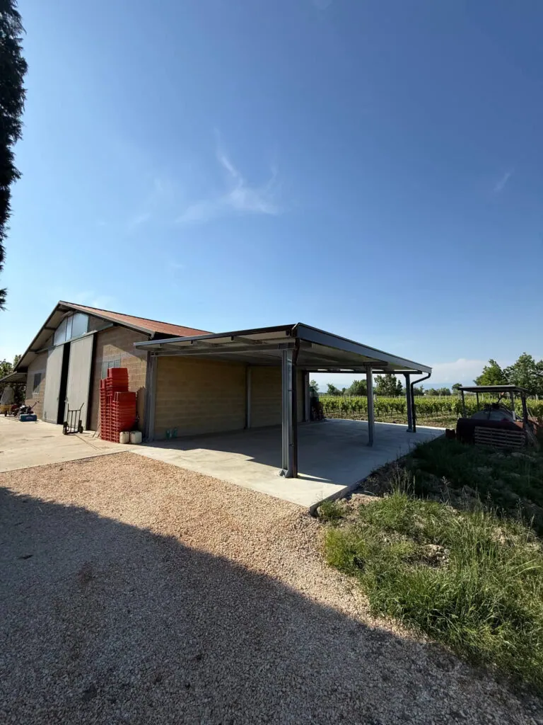 Grande tettoia metallica addossata a un edificio agricolo, realizzata con una solida struttura portante in acciaio e copertura in pannelli coibentati, adibita a rimessa per macchinari. Progettazione, realizzazione e posa a cura di Carpenteria 2A a Treviso, operante in tutto il Veneto - Vista 2 - foto tettoie in ferro.