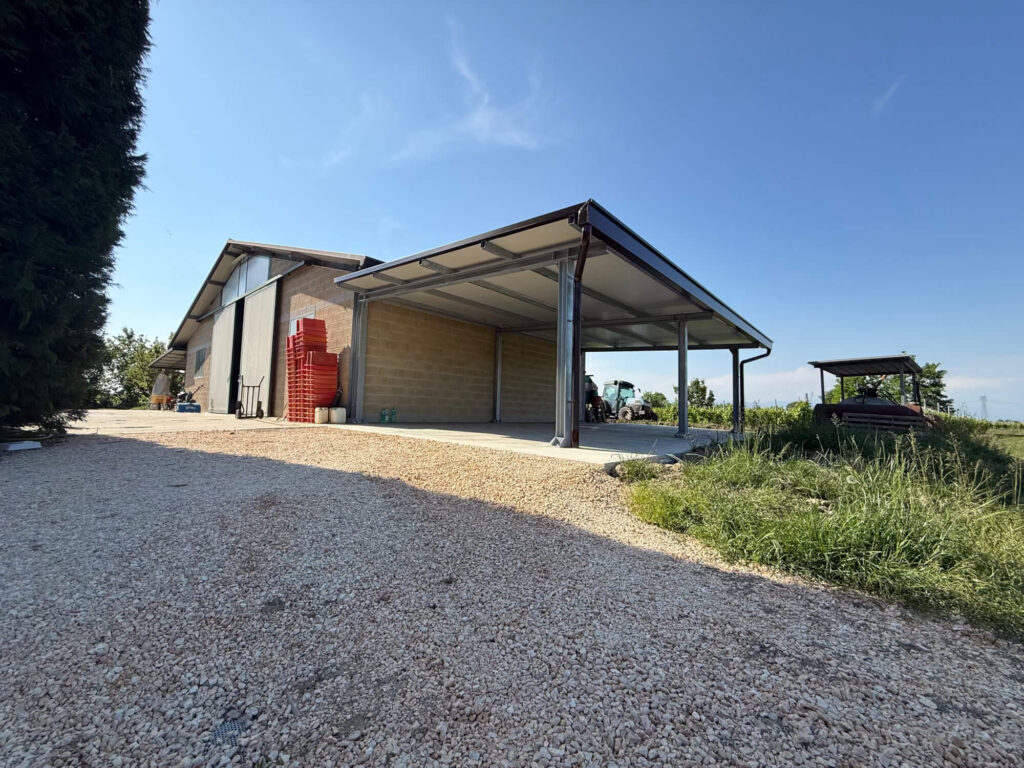 Grande tettoia metallica addossata a un edificio agricolo, realizzata con una solida struttura portante in acciaio e copertura in pannelli coibentati, adibita a rimessa per macchinari. Progettazione, realizzazione e posa a cura dell'azienda 2A a Treviso, operante in tutto il Veneto - Galleria carpenteria in ferro.